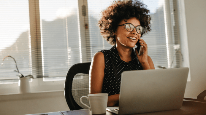 Lady talking on a cell phone, sitting at a desk, with her laptop open.