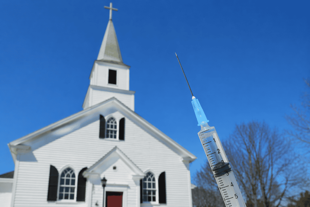 Photo of a traditional white chapel-style church with a steeple and cross in the background, slightly out of focus, set against a clear blue sky. In the foreground, a large, sharply focused vaccine syringe is angled upward, with visible measurement markings, creating a visual contrast between religious architecture and medical imagery.