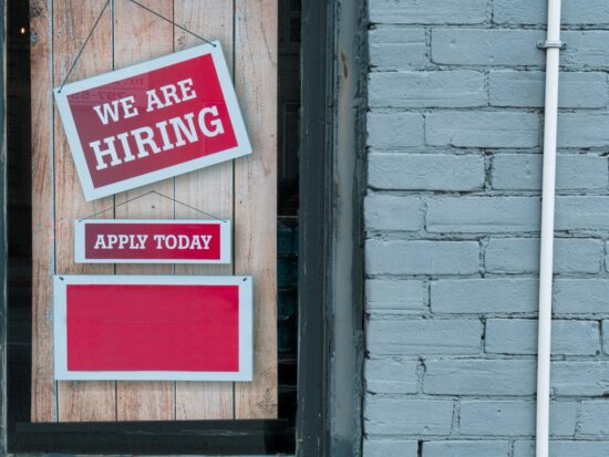 "We Are Hiring" Sign in Window of Brick Building - A red and white sign in a window of a brick building reads We Are Hiring with a smaller sign that says Apply Today.